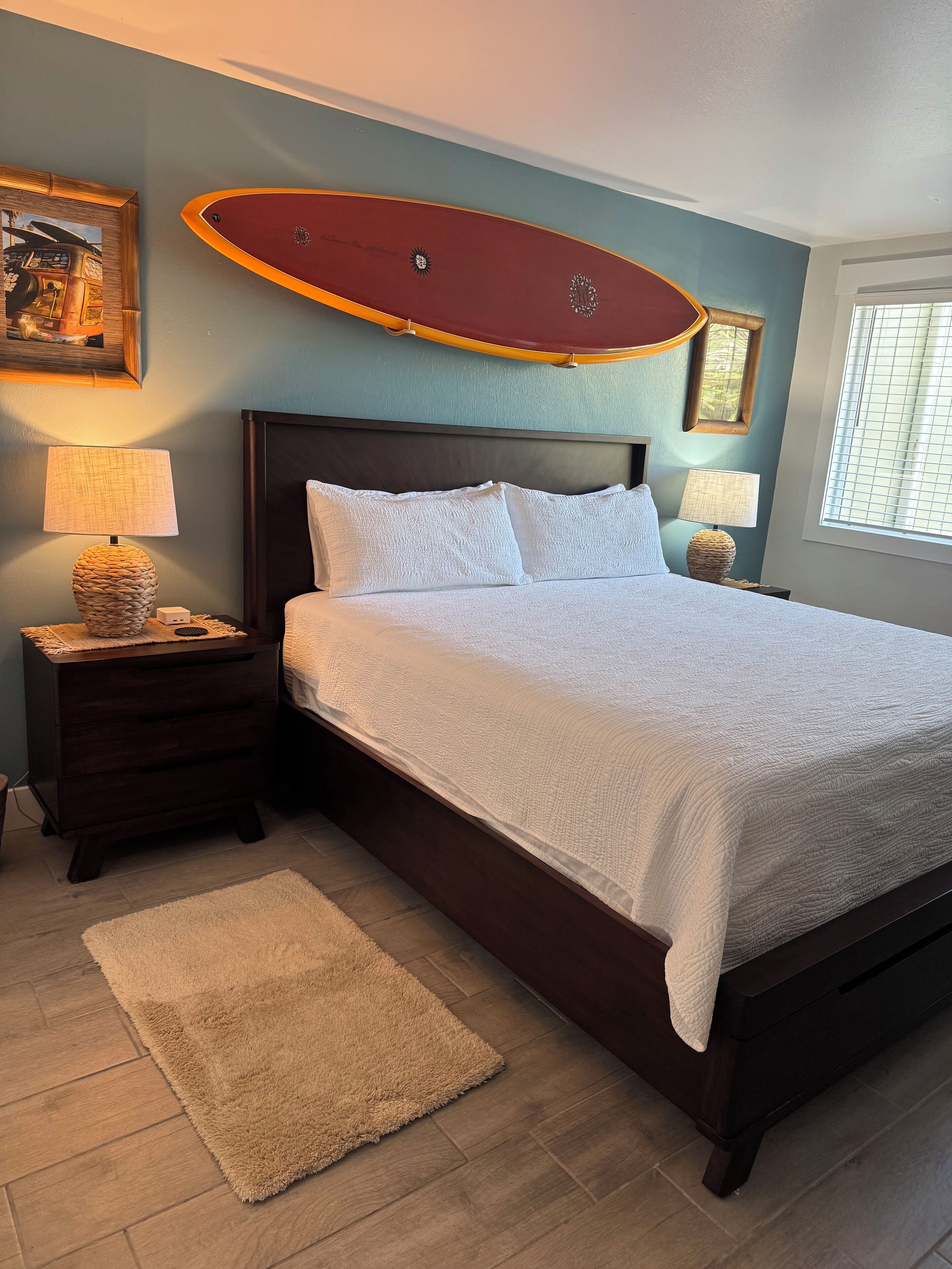 A bedroom with a large bed, two bedside tables with lamps, a wooden headboard, a framed picture on the wall, and a window with blinds.