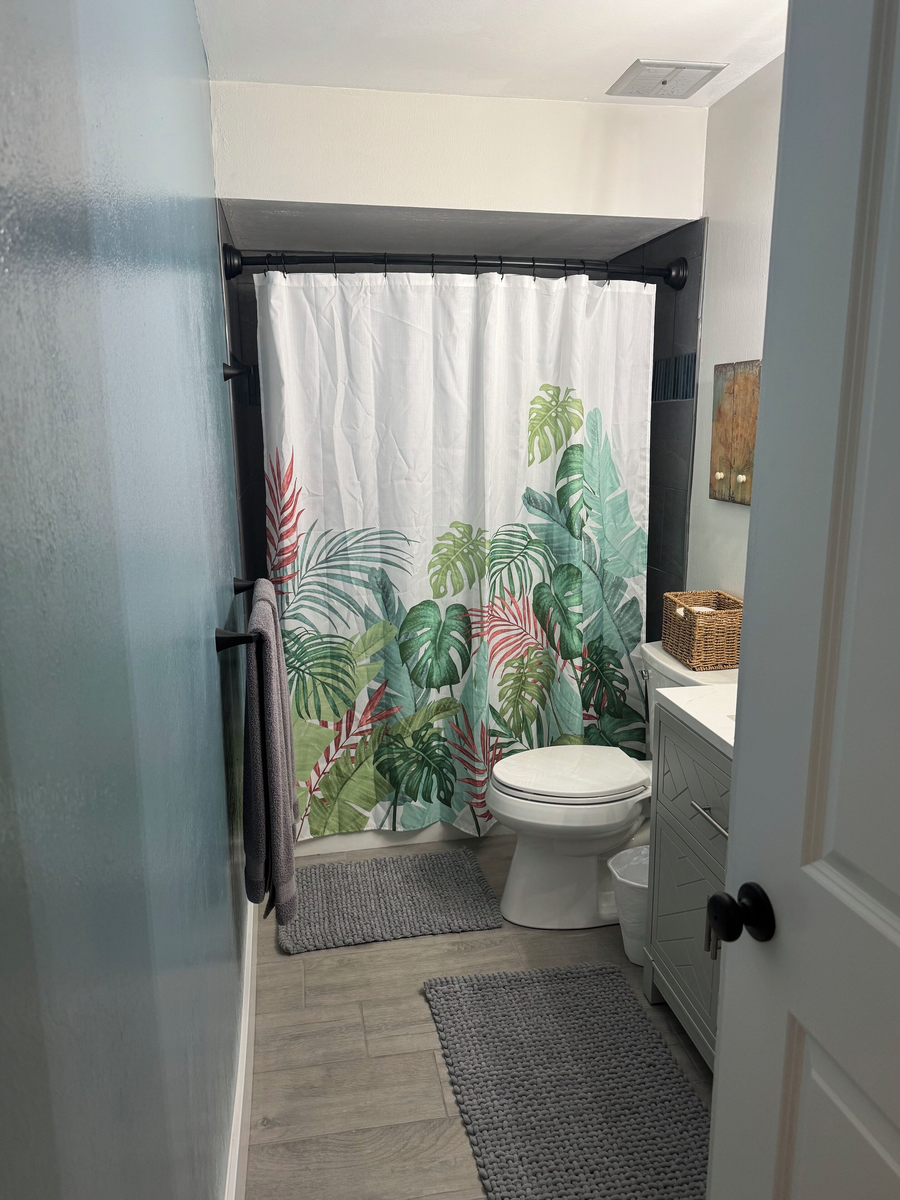 Ein Badezimmer mit einer Duschvorhang mit tropischem Blattmuster, einer Toilette, einem Waschbecken und einem kleinen Teppich.