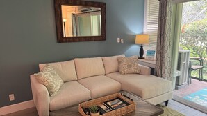 Una sala de estar con un sofá color beige, una mesa de centro con una bandeja, una lámpara y una ventana con cortinas.