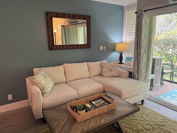 A living room with a beige sofa, a coffee table with a tray, a lamp, and a window with curtains. - Remodeled 2025, king bed, 2 Bathroom, sleeps 4 (Kahuku)