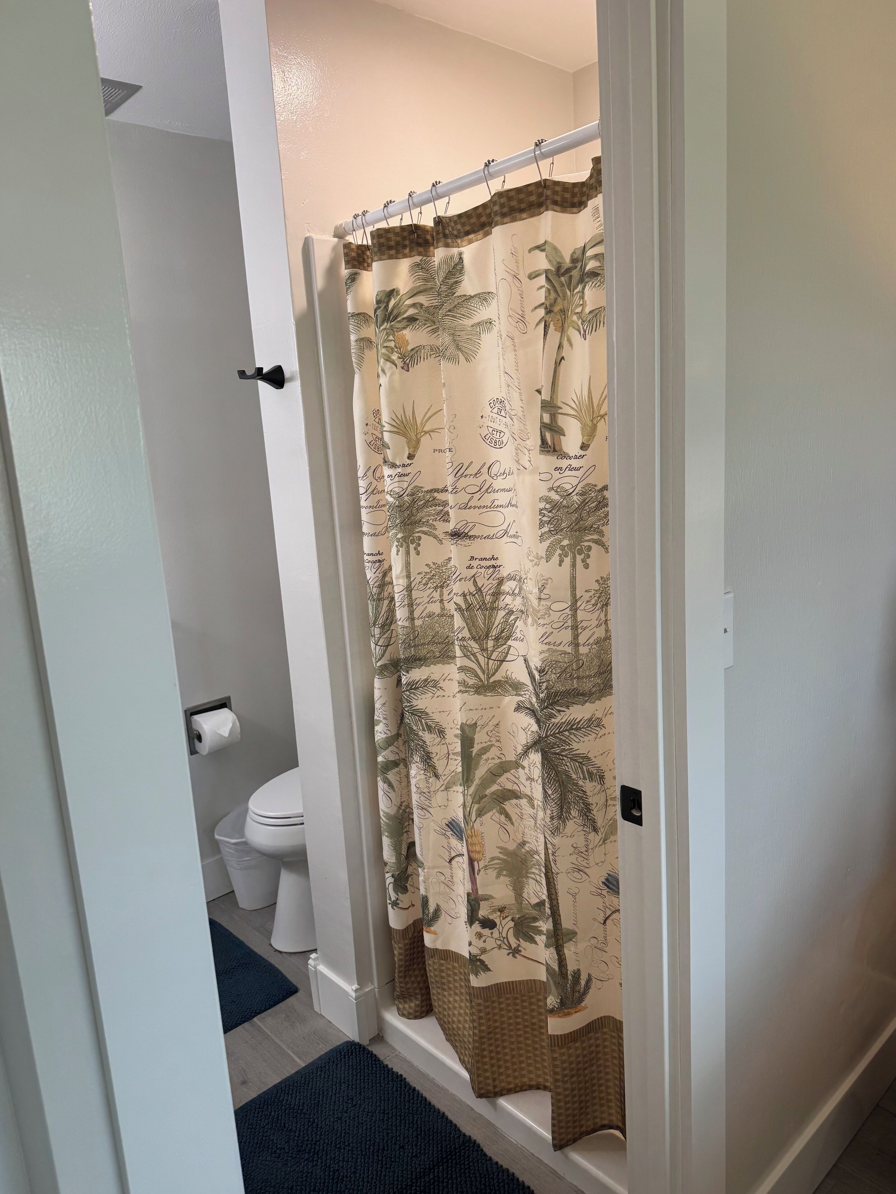 A bathroom with a shower curtain featuring palm tree and script patterns, a toilet, and a blue rug.