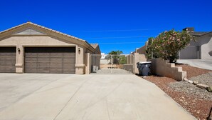Ein Auffahrtsweg führt zu einer Garage mit einem Metalltor, einem Betonweg und einer kleinen Gartenanlage mit einem Busch und einer Mülltonne.