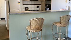 A modern kitchen with a central island, two wicker bar stools, and built-in appliances. - Riverside. Walk to Crown, CBD & MCG. Free Secure Carpark. (MELBOURNE)