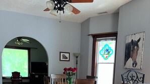 A dining area with a wooden table and chairs, a vase with red flowers, a ceiling fan, and a window with a horse painting.