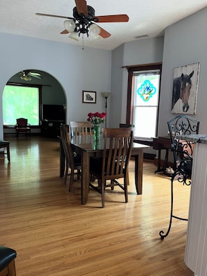 Un coin repas avec une table et des chaises en bois, un vase rempli de fleurs rouges, un ventilateur de plafond et une fenêtre ornée d’un tableau représentant un cheval.