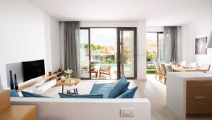A modern living room with a flat-screen TV, a sofa with blue cushions, a dining area with a table and chairs, and a balcony with a view of the city.
