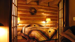 Ein gemütliches Schlafzimmer in einer Blockhütte mit einem Bett, das zwischen zwei Fenstern platziert ist, einer Holzbalkendecke und einem dekorativen Kranz an der Wand.