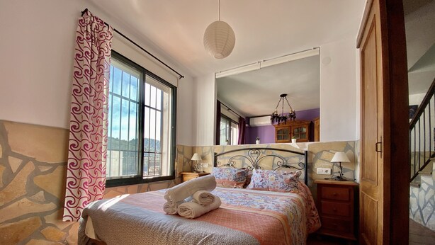 Un dormitorio con cama, ventana, espejo, mesita de noche y lámpara.