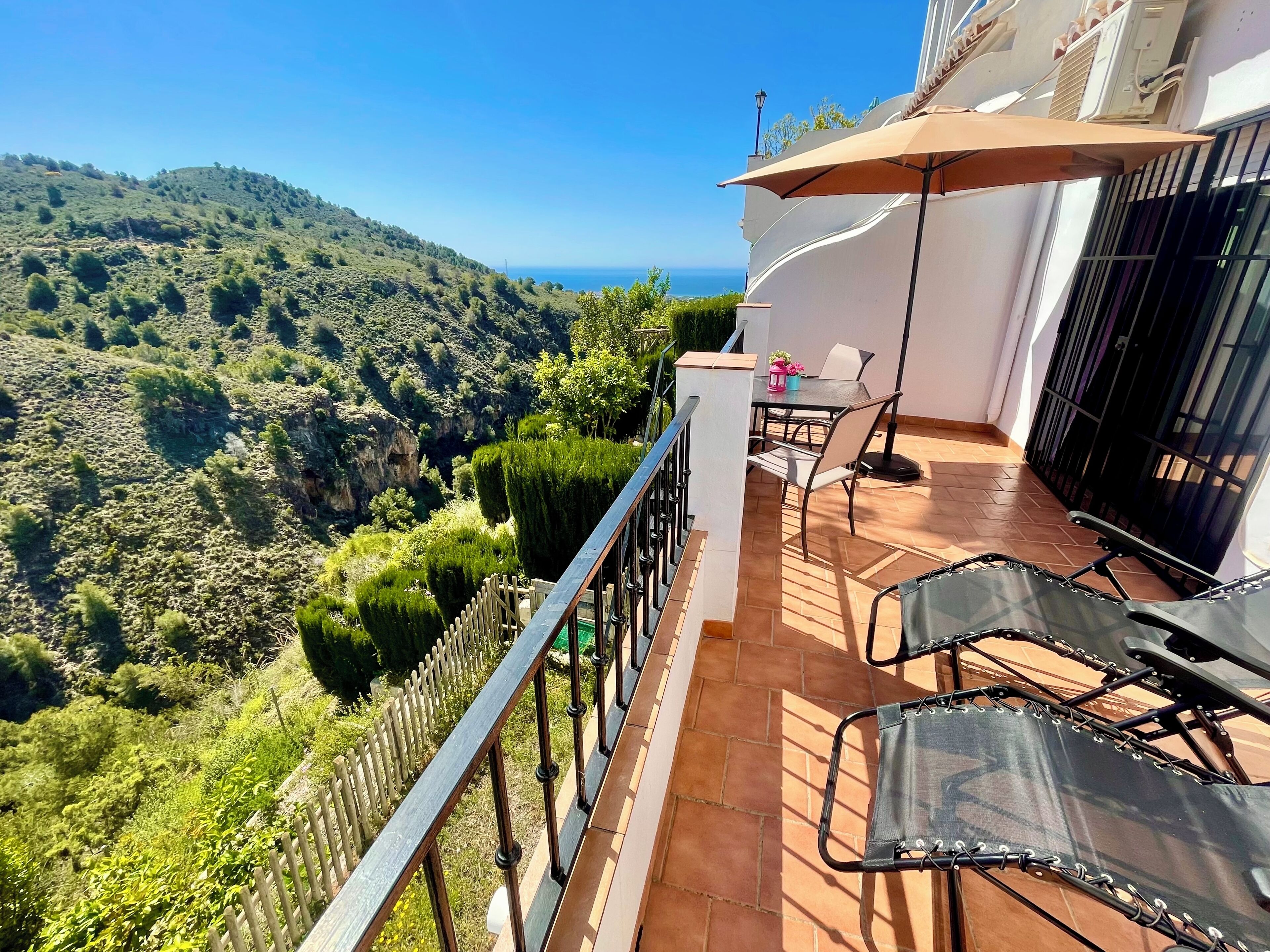 A balcony with a table and chairs, a view of the sea, and a mountainous landscape.