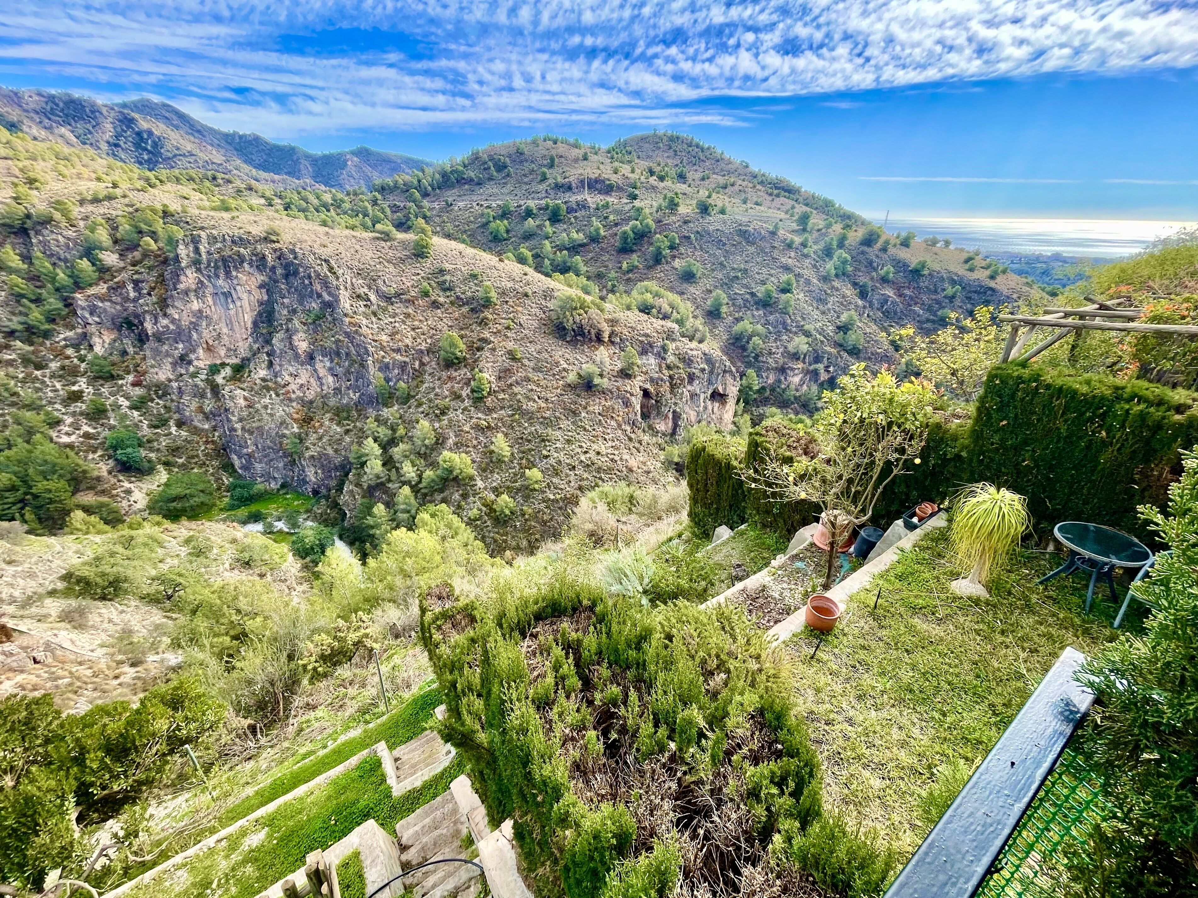 A mountainous landscape with a view of a valley and a distant body of water. There are terraced gardens and a small table with chairs.