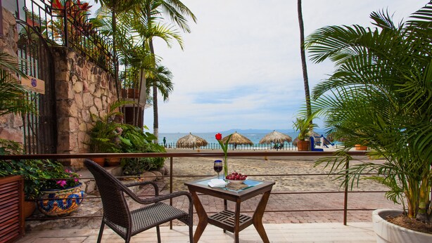 Ein Balkon mit Korbmöbeln, einem Tisch mit Weinglas und Obst, und Blick auf den Strand.