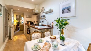 A cozy kitchen with a dining area, featuring a table set with tea cups, a bottle of champagne, and a loaf of bread. - New Church Cottage  - sleeps 4 guests  in 2 bedrooms (Tetbury)