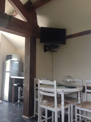 A dining area with a round table, white chairs, a television mounted on the wall, and a microwave in the background. - Cosy Chalet - Sleeps 2-3,  Peaceful Setting, 5kms To Shops,Linen/Towels Included (Romagny Fontenay)