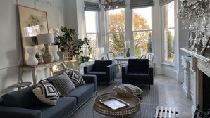 A spacious living room with a dark sofa, armchairs, a coffee table, and a fireplace. Large windows offer views of trees and a white exterior wall. - High View Large Luxury House (London)