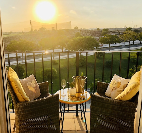 A balcony with wicker chairs, a small table with a bottle and glasses, and a view of a road and greenery.