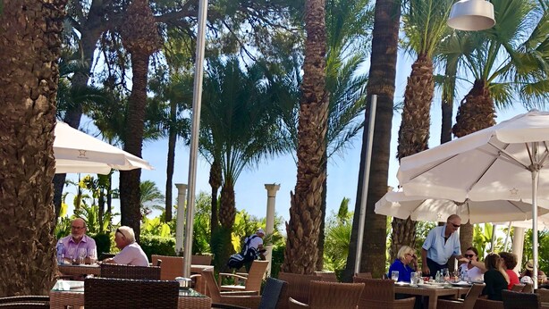 An outdoor dining area with wicker furniture and umbrellas, surrounded by palm trees.
