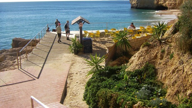 A coastal walkway leading to a beach with yellow chairs and tables, a signpost with a no swimming symbol, and a rocky cliff in the background. - Apartment Tropicos (Carvoeiro)