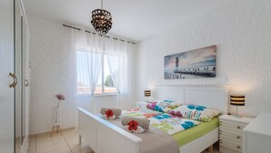 A bedroom with a bed, bedside lamps, a window with sheer curtains, a painting on the wall, and a small table with a vase. - Apartment Tropicos (Carvoeiro)