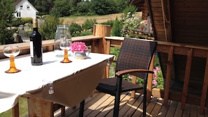 Terraza de madera al aire libre con una mesa lista para comer, rodeada de vegetación y una casa al fondo.