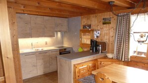 L’intérieur d’un chalet en bois comprend une cuisine équipée d’un évier, d’une cuisinière et d’une machine à café. On y trouve une table à manger avec des chaises, ainsi qu’un petit îlot central avec un banc.