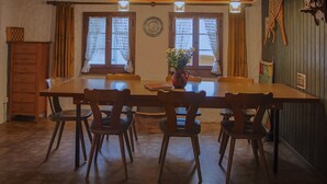 A dining area with a long wooden table, six chairs, two windows with lace curtains, a vase with flowers, and a wooden cabinet. - House in the middle of the Swiss Alps (Wassen)