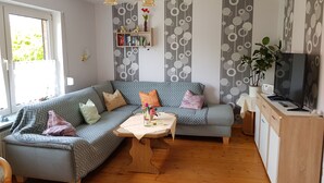 Ein modernes Wohnzimmer mit einer Couch, einem Couchtisch und einer TV-Konsole. Ein Bücherregal beherbergt Bücher und Dekoration, und ein Fenster bietet Blick auf Grünflächen.