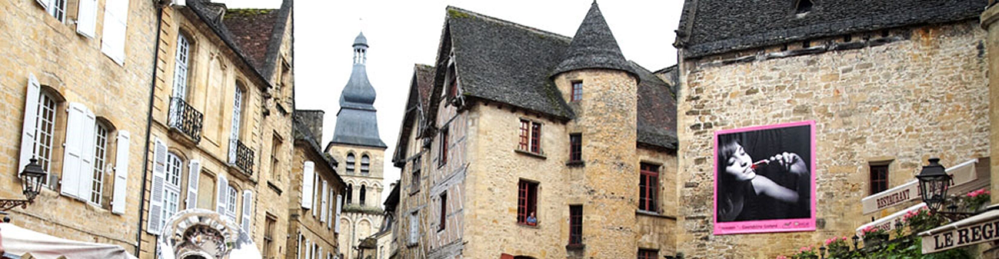 A narrow street lined with historic stone buildings, featuring a prominent tower and a sign for 'LE REGIS'.