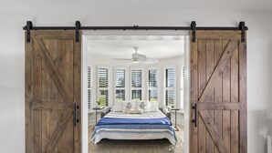 Ein Schlafzimmer mit einem Bett, einem Deckenventilator und Blick durch offene Holztüren in einen Raum mit Fenstern und Grünflächen.