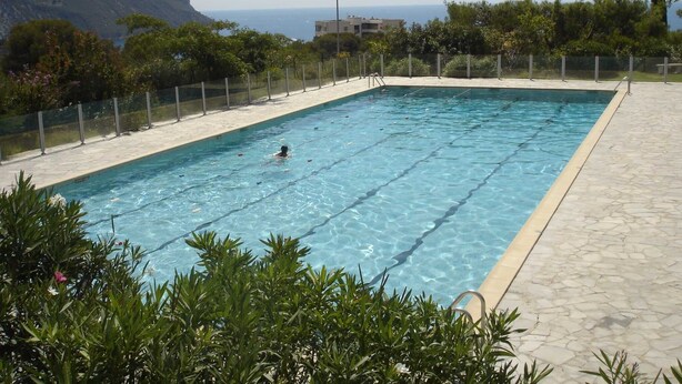 Ein rechteckiger Swimmingpool mit klarem, blauem Wasser, umgeben von einer Steindeck und einem Metallzaun. Im Hintergrund sind ein Felsen und ein Gebäude zu sehen.