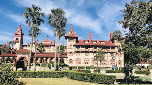 Un gran edificio histórico con múltiples torres y arcos, rodeado de jardines cuidados y palmeras.