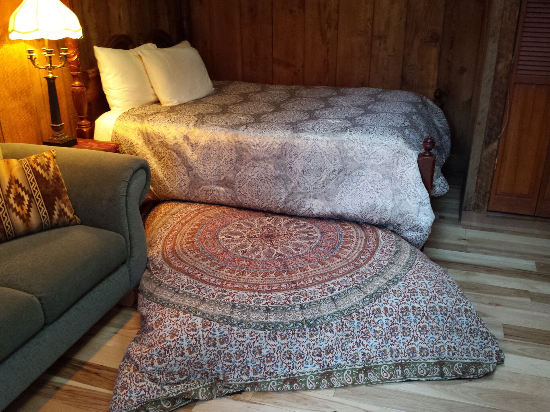 A room with a bed covered in patterned bedding, a grey sofa with a patterned throw, and a wooden floor.
