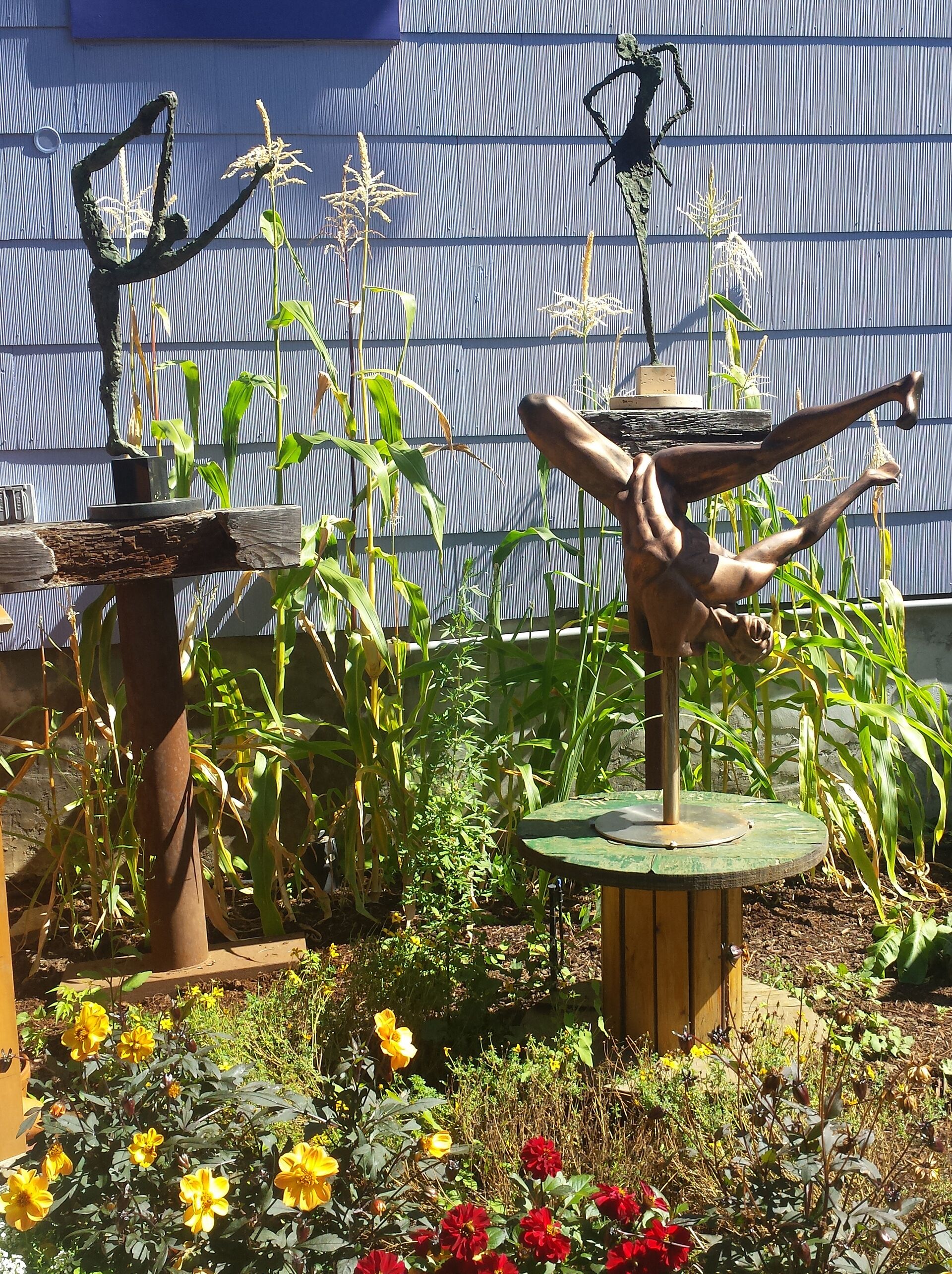 Un giardino con sculture, piante di mais e fiori.