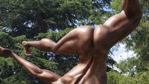 Escultura de bronze de uma figura humana em pose dinâmica, situada num jardim com cerca de madeira, um banco e vegetação ao fundo.