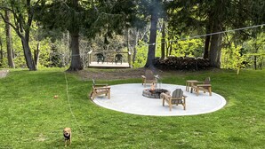 Un espace extérieur circulaire aménagé avec des sièges, doté d’un foyer, entouré de chaises et de tables en bois, et situé dans une cour verdoyante avec des arbres en arrière-plan.