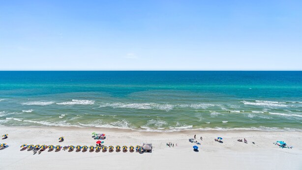 Ein Strand mit Reihen von Sonnenschirmen und Liegestühlen, einem klaren blauen Himmel und ruhigen Wellen.