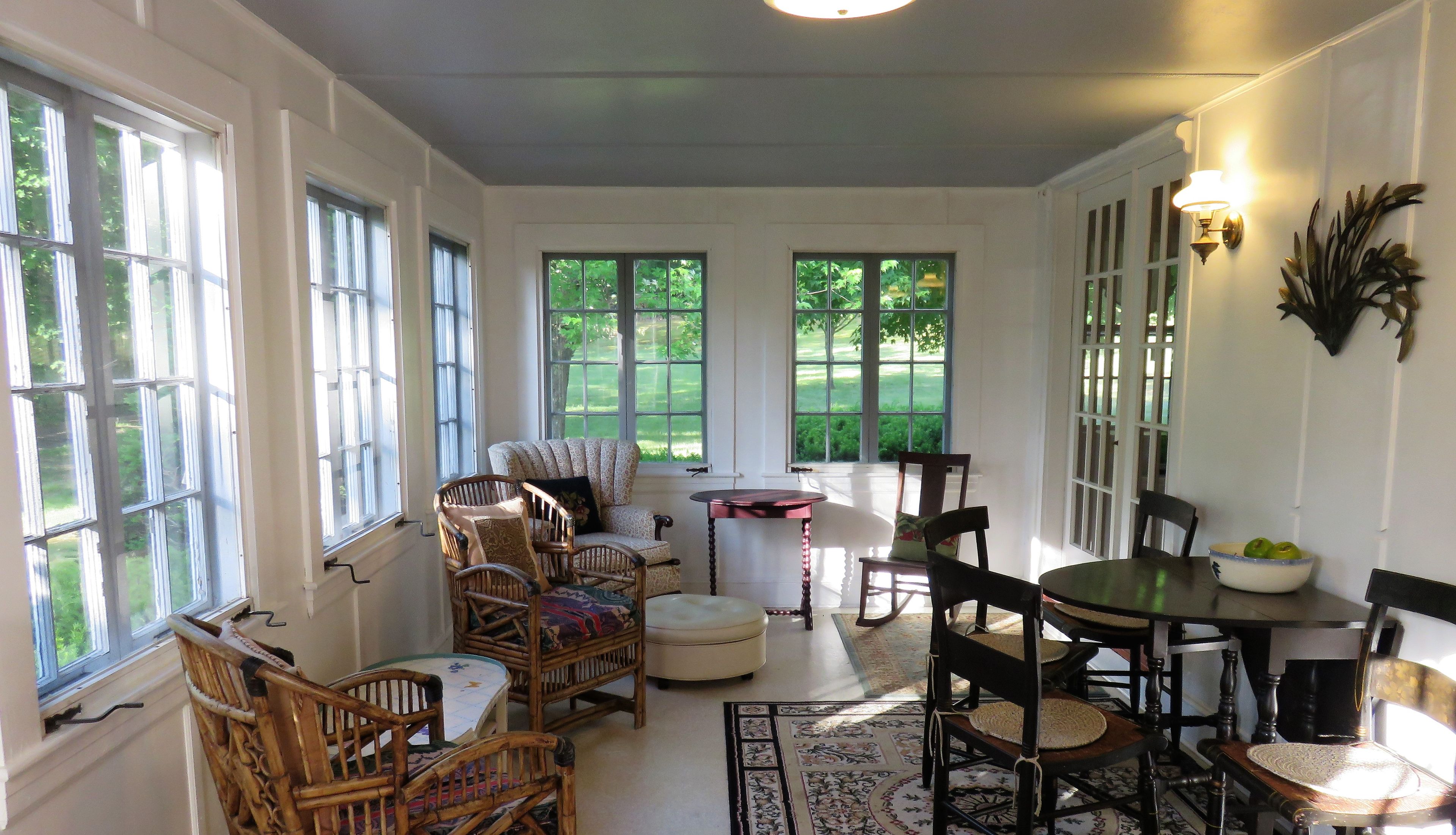 A well-lit room with large windows, wicker furniture, a round table with chairs, and a decorative wall piece.