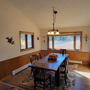 A dining room with a wooden table, chairs, and a chandelier. There are windows with curtains and a view of trees outside.
