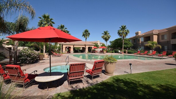 A pool area with red chairs and umbrellas, surrounded by palm trees and a building with multiple balconies.