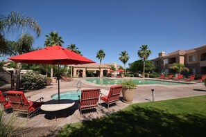 A pool area with red chairs and umbrellas, surrounded by palm trees and a building with multiple balconies.