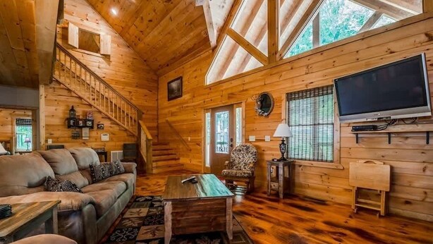A wooden cabin interior with a sofa, coffee table, TV, and a staircase.