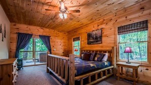 A log cabin bedroom with a bed, a ceiling fan, a window with a view of trees, and a small table with a lamp.