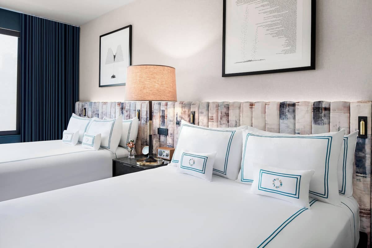 Une chambre d’hôtel avec deux lits, une grande fenêtre avec des rideaux bleus, une lampe fixée au mur et des œuvres d’art encadrées.