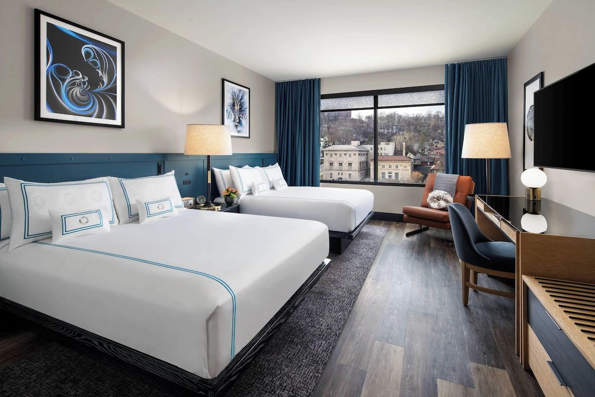 Une chambre d’hôtel avec deux lits, un bureau, une chaise, une télévision et une fenêtre donnant sur des immeubles.
