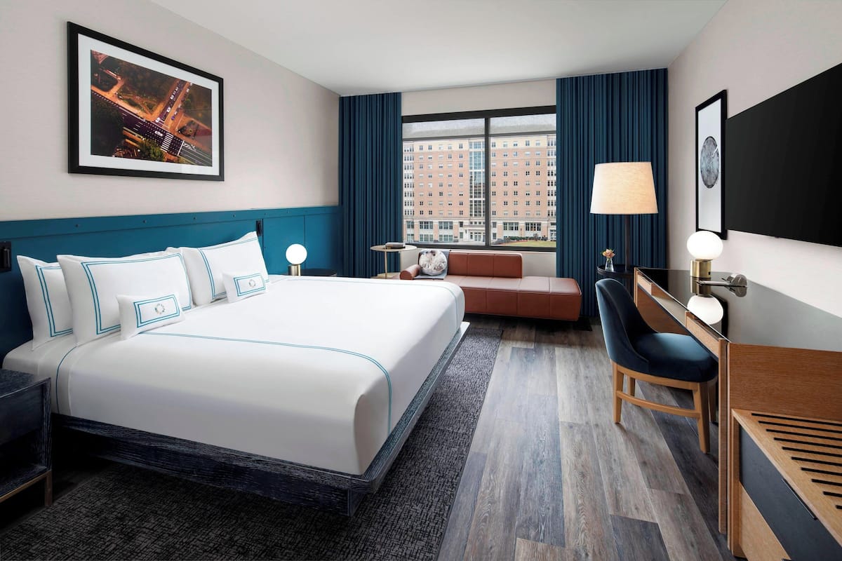 Une chambre d’hôtel avec un grand lit, un bureau, une chaise, un canapé, une lampe et une vue sur les immeubles par la fenêtre.