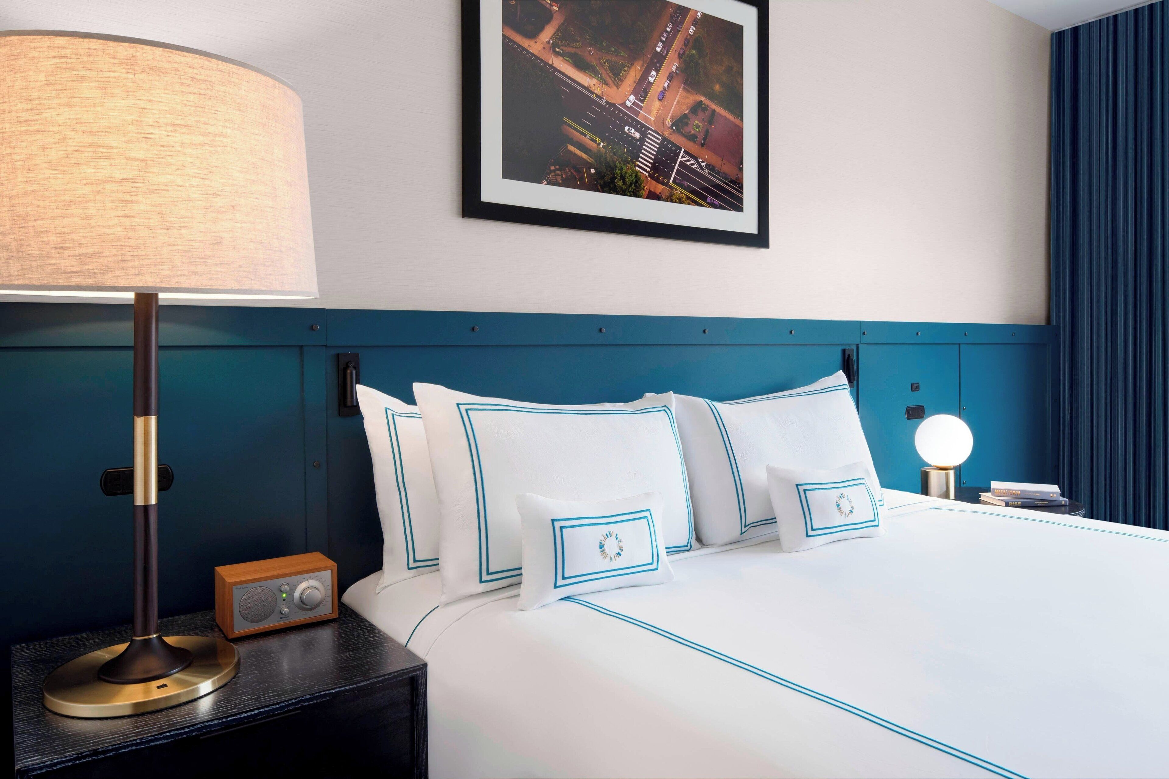 Un lit bien fait, avec du linge de lit blanc et des bordures bleues, une table de chevet avec une lampe et une radio, et un tableau représentant un paysage urbain encadré accroché au mur.