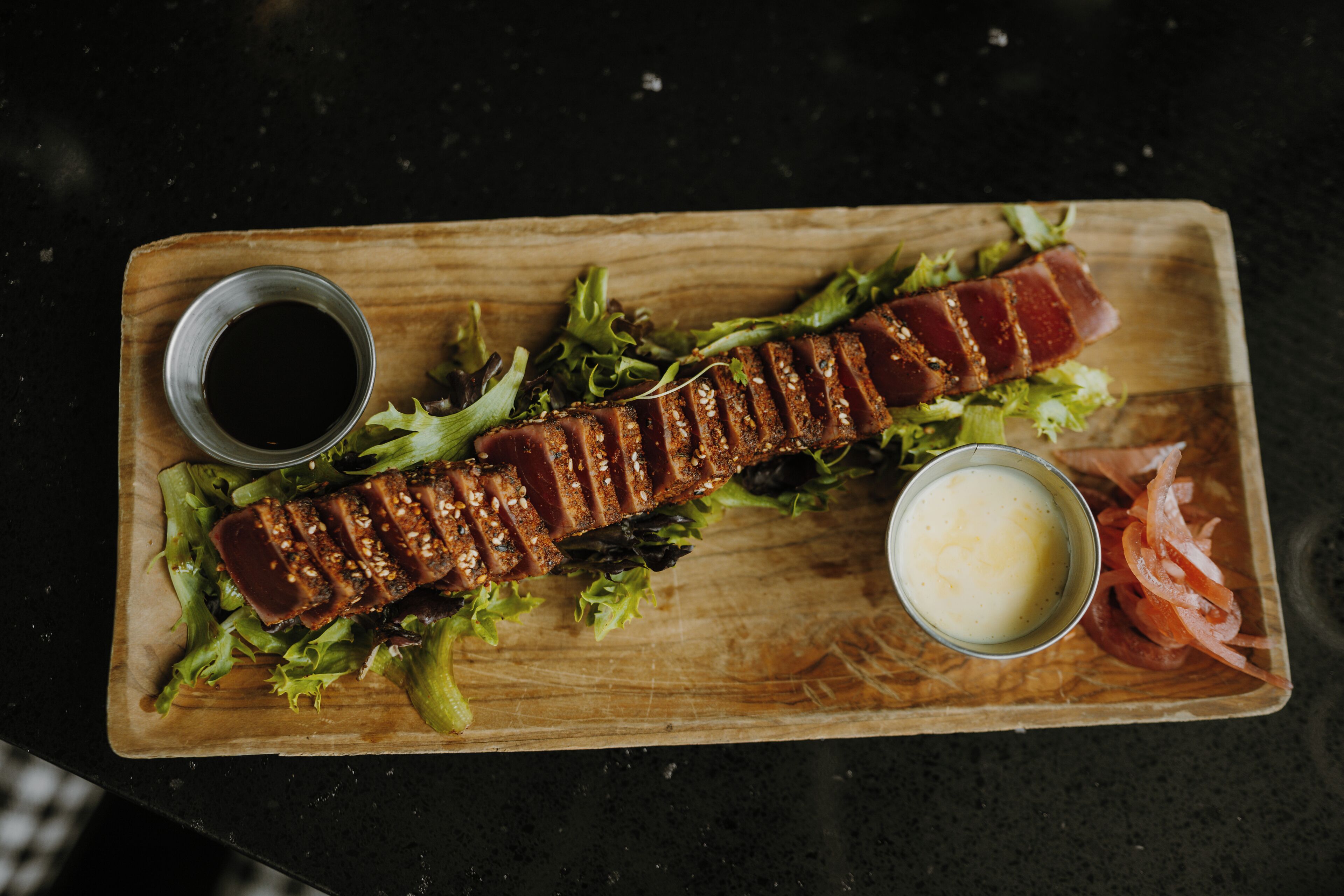 A wooden serving board with a grilled meat dish, greens, and two small bowls of sauce.