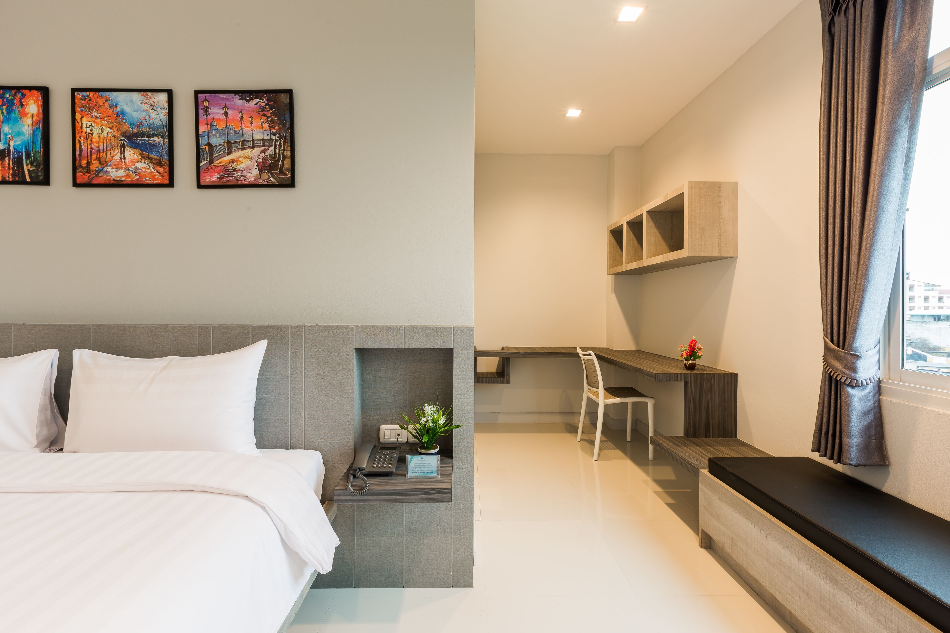 A modern hotel room with a bed, a desk, and a seating area. There are three paintings on the wall.