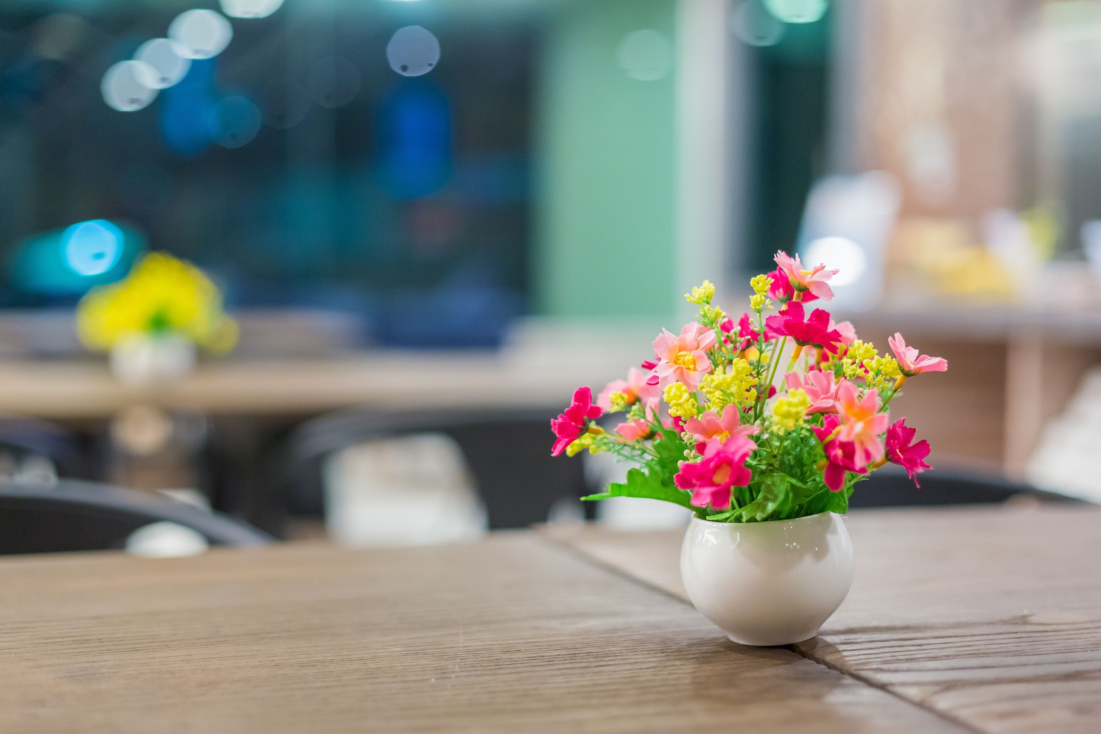 A white vase with pink and yellow flowers on a wooden table in a room with a blurred background.