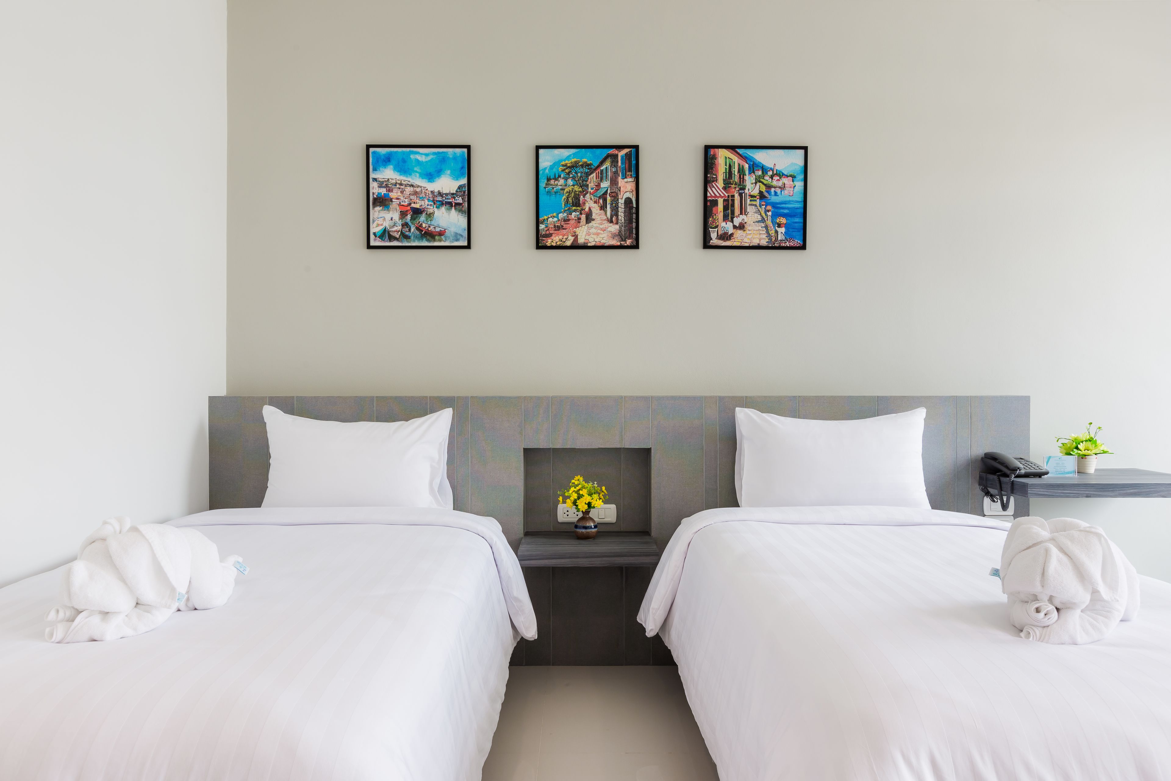 A hotel room with two beds, a nightstand with a vase of flowers, and three framed pictures on the wall.