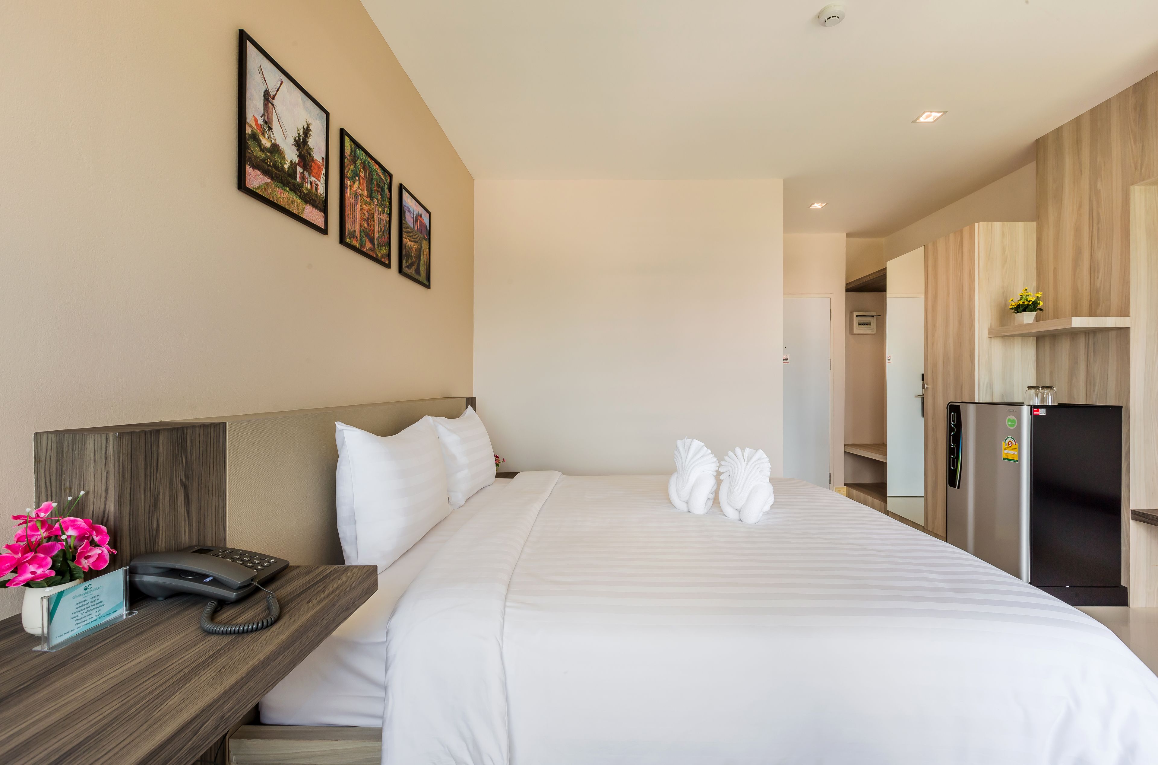 A hotel room with a bed, a desk with a phone, a refrigerator, and a wall with framed pictures.